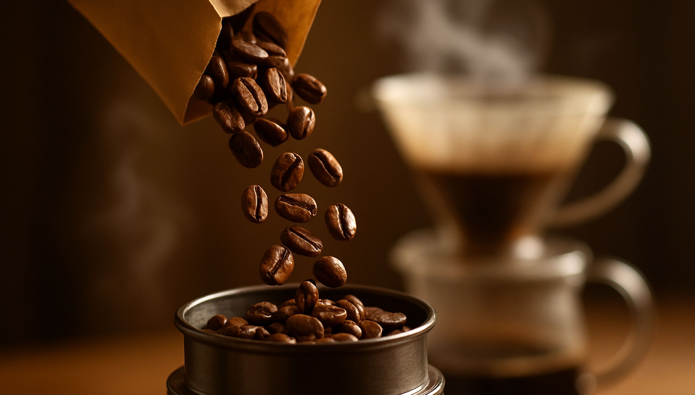 A close-up of roasted coffee beans being poured into a grinder or stored in a glass jar. Light steam rises in the background from a brewing coffee setup.